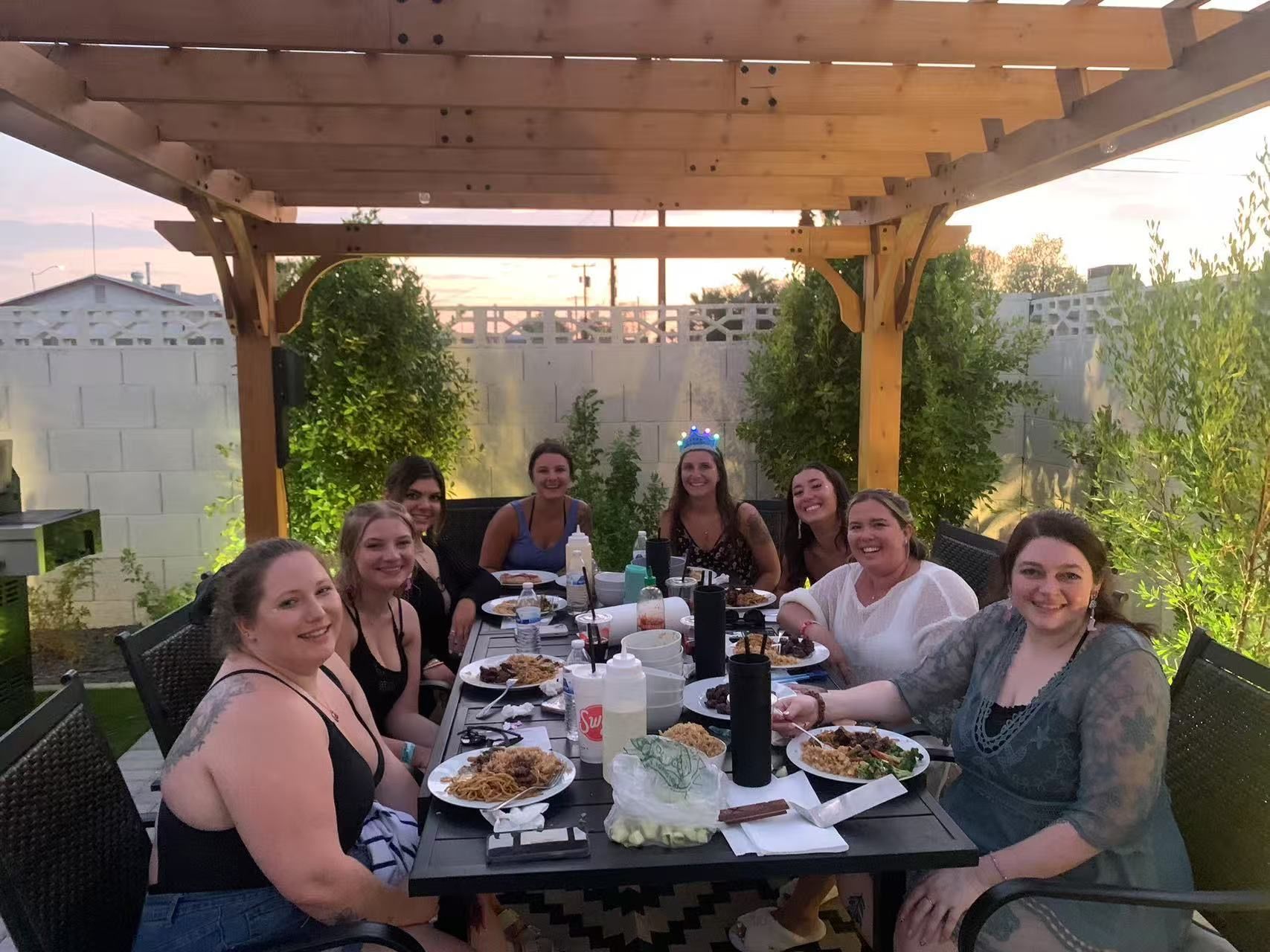 Group of friends enjoying an evening backyard patio dinner under a wooden pergola at sunset, plates of food on the table and one guest wearing a light-up party crown.