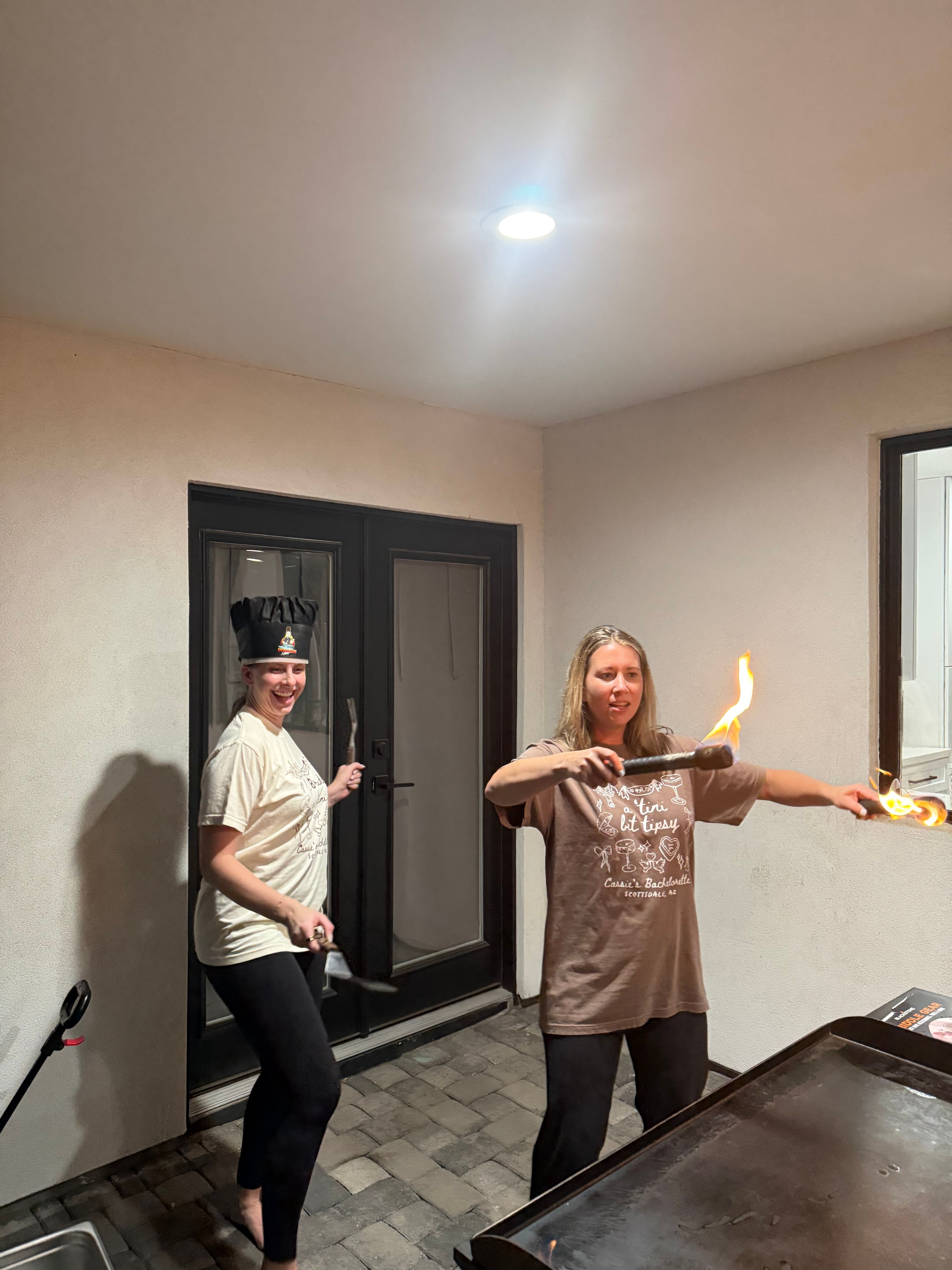 Two people on a home patio griddle at night — one wearing a chef hat and holding a spatula, the other waving two flaming torches over a flat-top grill beneath a recessed ceiling light on stone pavers.