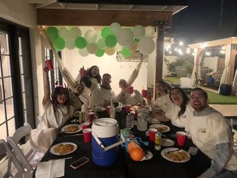 Friends cheering at a nighttime backyard patio party under a wooden pergola with green-and-white balloons and string lights, gathered around a long table with plates of pasta, red cups, a blue cooler and a water toy.