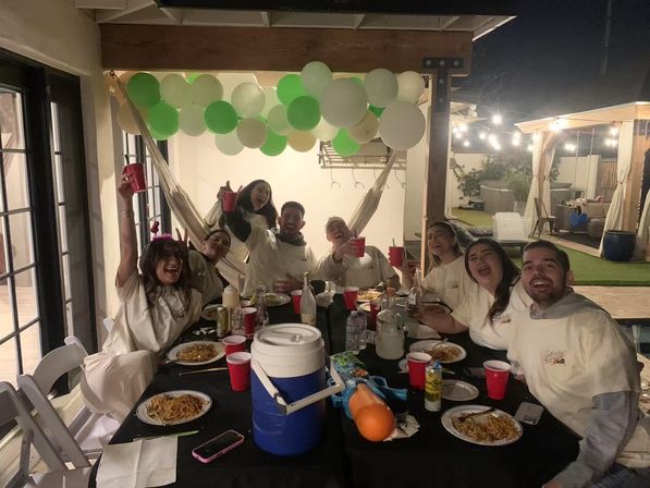 Friends cheering at a nighttime backyard patio party under a wooden pergola with green-and-white balloons and string lights, gathered around a long table with plates of pasta, red cups, a blue cooler and a water toy.