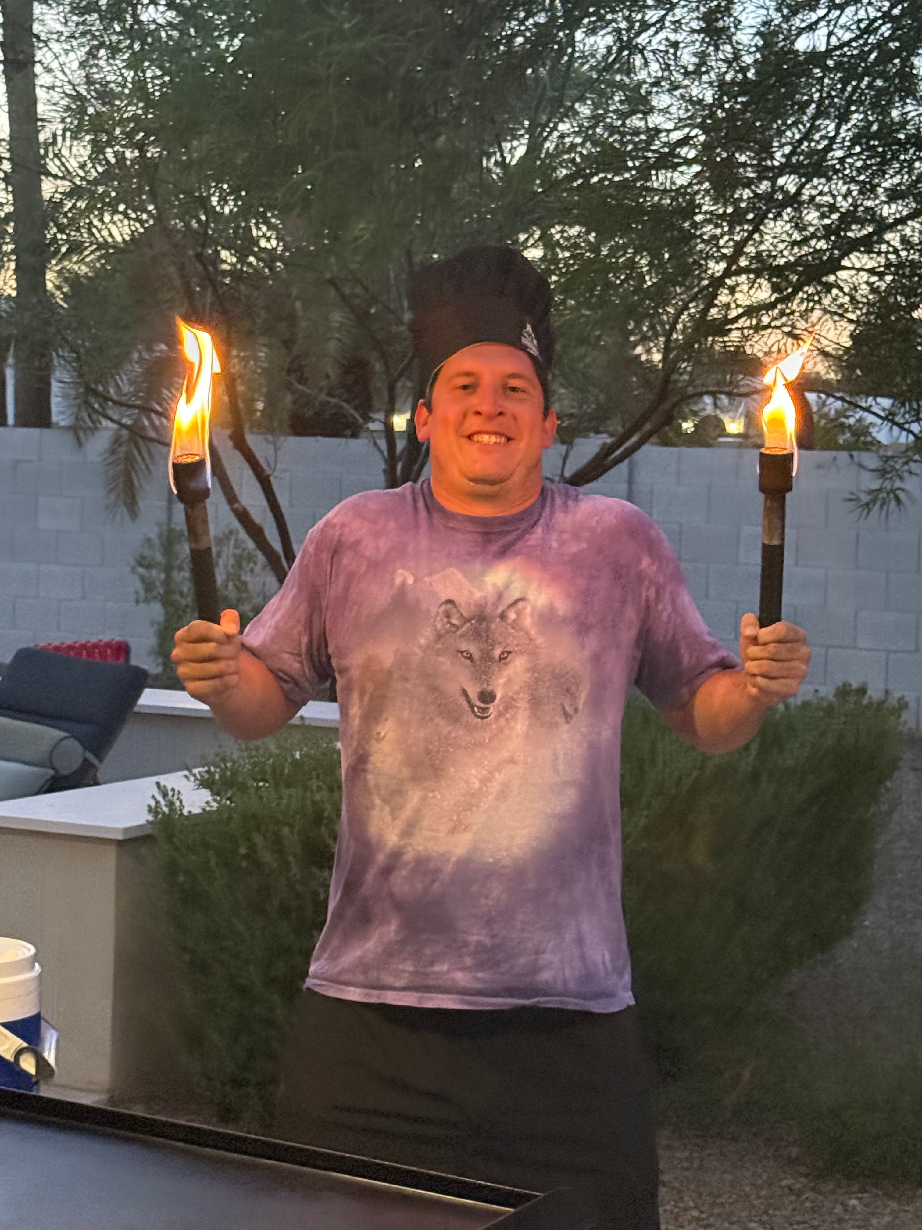 Person wearing a black hat and purple wolf T‑shirt holding two lit tiki torches aloft in a dusk backyard with desert plants and a cinderblock wall