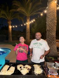 Two people smiling by a lit backyard pool at night under palm trees and string lights, grilling rice shaped into a heart and letters on a flat-top griddle.
