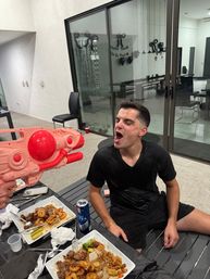 Young man on a modern patio with mouth open as a pink toy water blaster sprays him, table with plates of shrimp and steak, a beer can, and sliding glass doors in the background