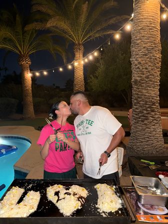 Couple kissing while outdoor grilling by a backyard pool at night, string lights and palm trees above, rice arranged into a 'U ♥ I' on the griddle.