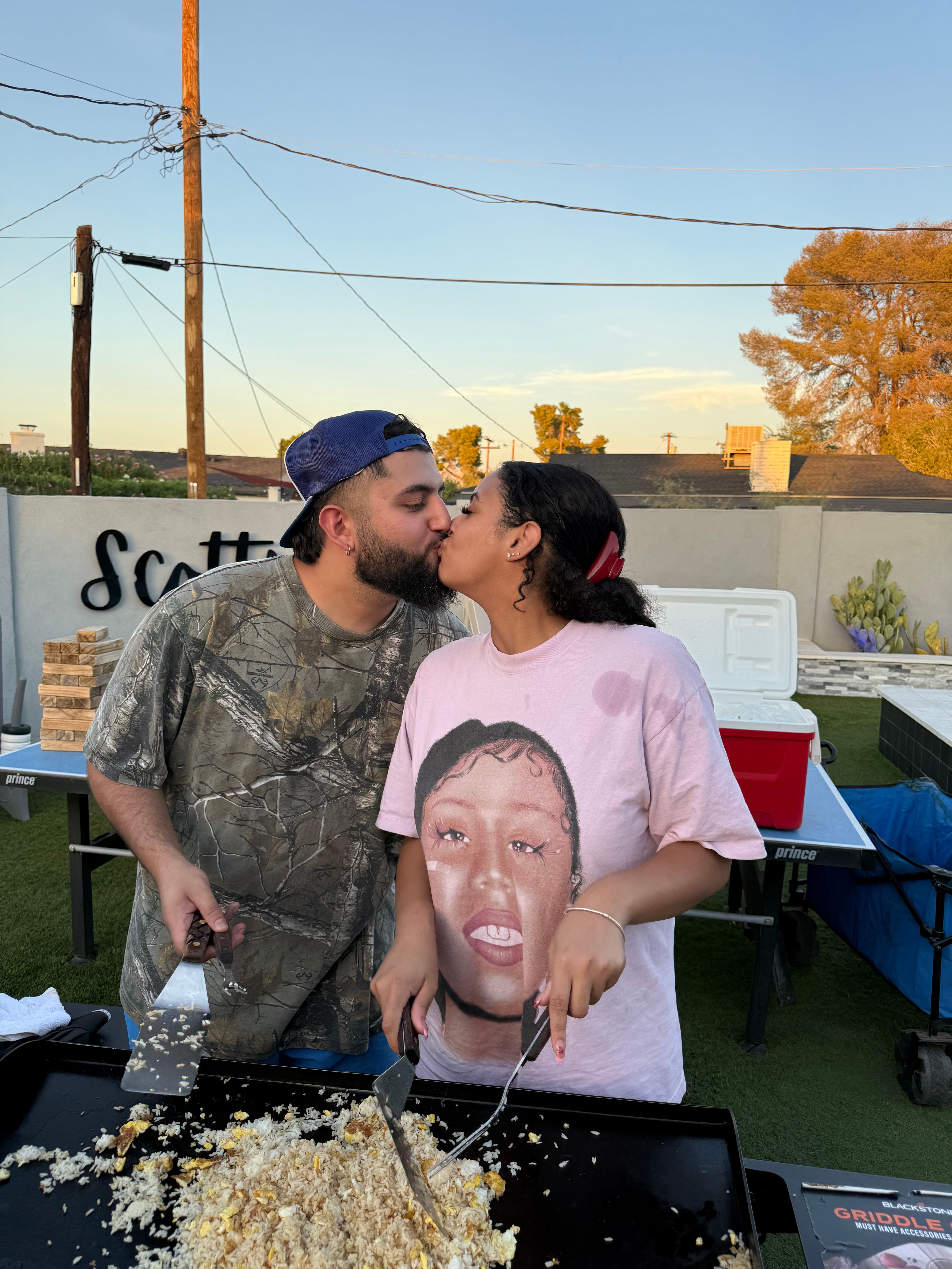Couple kissing over an outdoor griddle in a backyard at sunset, flipping fried rice and eggs with spatulas; man in camo shirt and backward cap and woman in a pink graphic tee with a cooler and patio gear in the background.