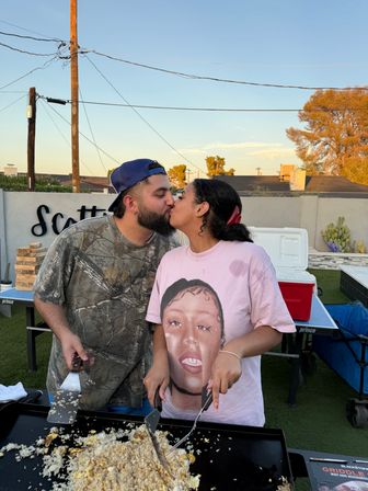 Couple kissing over an outdoor griddle in a backyard at sunset, flipping fried rice and eggs with spatulas; man in camo shirt and backward cap and woman in a pink graphic tee with a cooler and patio gear in the background.