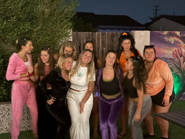 Group of young women posing and laughing at a backyard night party, wearing colorful casual outfits in front of a wooden fence and painted mural.