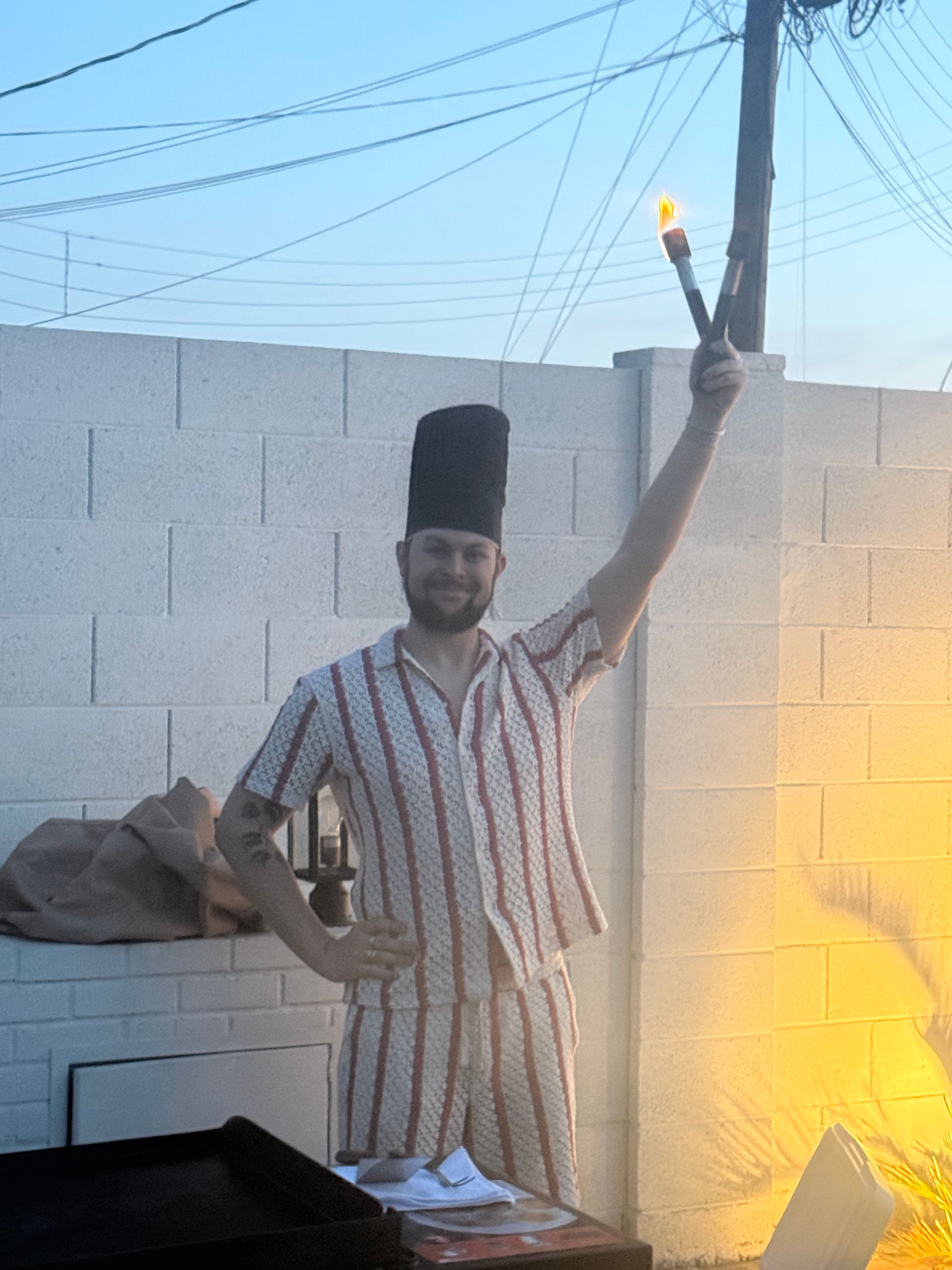 Person in a tall chef’s hat and red-striped matching shirt and shorts holding a lit torch high in a backyard at dusk, grill and plates on a table, cinderblock wall and power lines in background, warm yellow light on one side