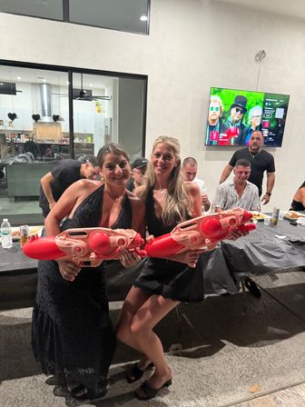 Two women in black dresses strike a playful pose on an outdoor patio at night, each holding oversized red water blasters, with a buffet table, seated guests and a wall-mounted TV visible in the background.