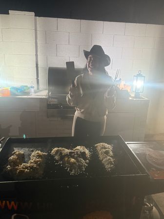 Person in a cowboy hat holding a spatula and tongs at a backyard flat-top griddle at night, cooking rice under lantern light on an outdoor patio