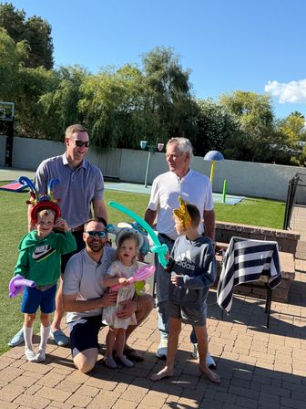 Multigenerational family enjoying a sunny backyard with artificial turf, kids wearing colorful balloon hats and holding balloon swords while adults smile on the patio.