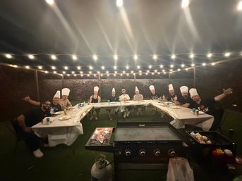 Backyard patio dinner at night under string lights — a group of adults in chef hats seated around U-shaped tables with plates and drinks and a large flat-top griddle in the foreground.