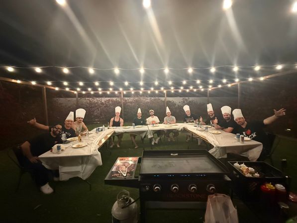 Backyard patio dinner at night under string lights — a group of adults in chef hats seated around U-shaped tables with plates and drinks and a large flat-top griddle in the foreground.