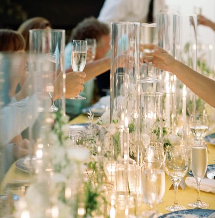 Guests clinking champagne flutes over an elegant wedding reception table with tall glass candle vases, a floral runner, and warm candlelight