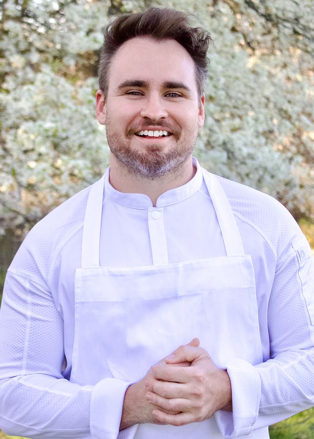 Smiling chef in a white jacket and apron, hands clasped, standing outdoors with spring blossoms in the background.