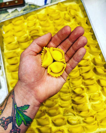 Tattooed forearm holding three bright yellow handmade tortellini over a tray of neatly arranged fresh stuffed pasta.