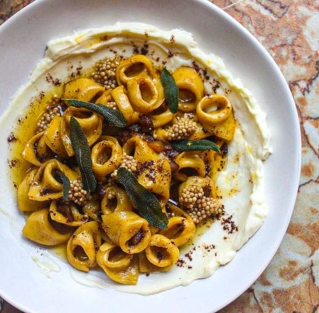 Overhead shot of pillowy tortellini in brown-butter and sage, served on a creamy ricotta smear and dotted with whole mustard seeds on a white plate over a stone tabletop.