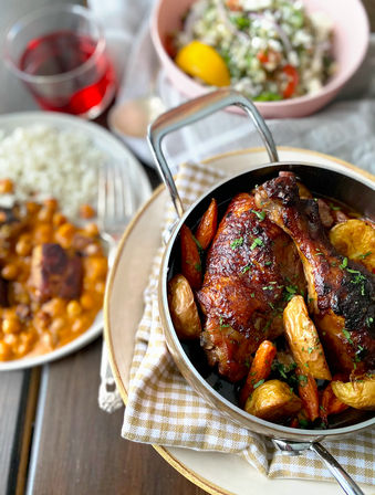 Mouthwatering roasted chicken legs with golden baby potatoes and glazed carrots in a skillet on a gingham napkin, paired with rice and chickpea stew, a fresh lemony salad and a red drink on a wooden table.