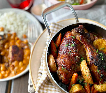 Mouthwatering roasted chicken legs with golden baby potatoes and glazed carrots in a skillet on a gingham napkin, paired with rice and chickpea stew, a fresh lemony salad and a red drink on a wooden table.