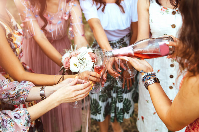 Friends in floral dresses toasting with rosé champagne flutes outdoors as one pours from a bottle, bouquet of pink-and-white roses, summer celebration vibe