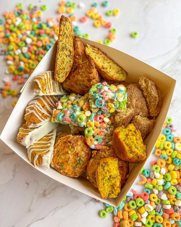Assorted breakfast pastry box with golden scones, glazed cake slices, cinnamon-sugar biscuits and colorful cereal-loop clusters on a marble countertop