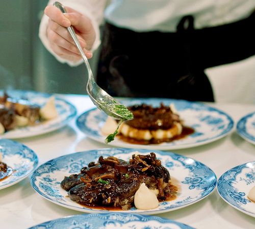 Server spooning bright herb garnish over plated braised short ribs with mushroom sauce and pearl onions on blue floral china in a fine-dining restaurant kitchen