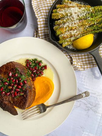 Seared steak topped with pomegranate-herb salsa and sweet potato puree on a white plate with a fork; parmesan-crusted roasted asparagus and lemon in a cast-iron skillet, glass of red wine on a marble tabletop.