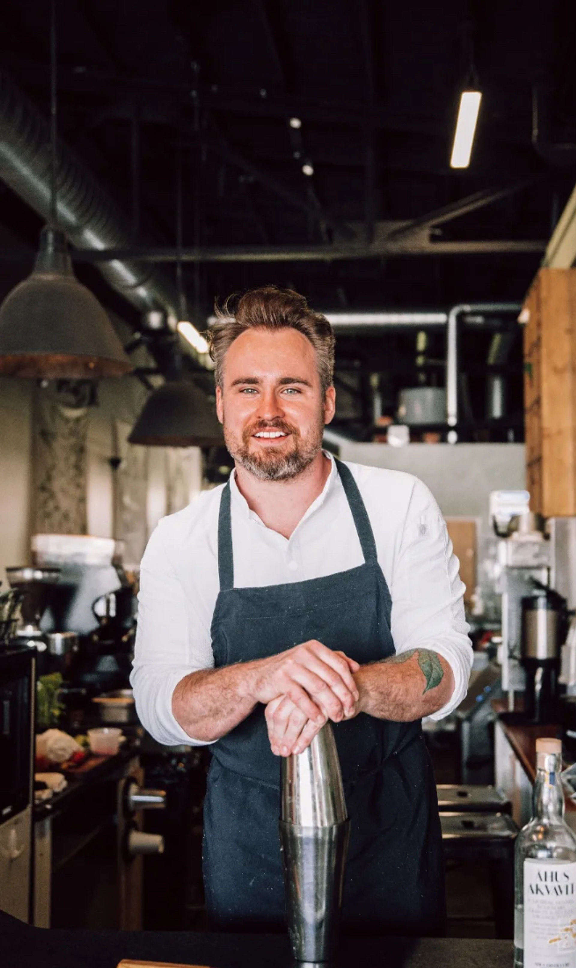 Smiling barista in white shirt and dark apron leaning on a metal cocktail shaker at an industrial-style café counter, with exposed ductwork, a chef’s knife on the bar and a bottle nearby.