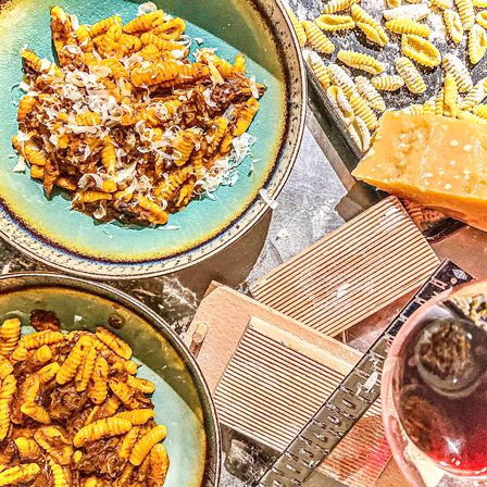 Two turquoise bowls of ridged cavatelli pasta tossed in a rich meat ragu and shaved Parmesan, with a tray of fresh pasta, a block of Parmesan and grater, and a glass of red wine on a marble countertop.