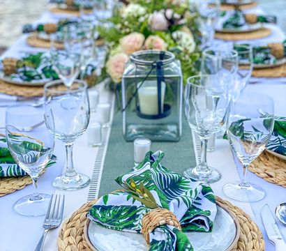 Al fresco waterfront tablescape with woven chargers, white plates, tropical leaf napkins in rattan rings, clear wine glasses, a glass lantern candle and a lush floral centerpiece.