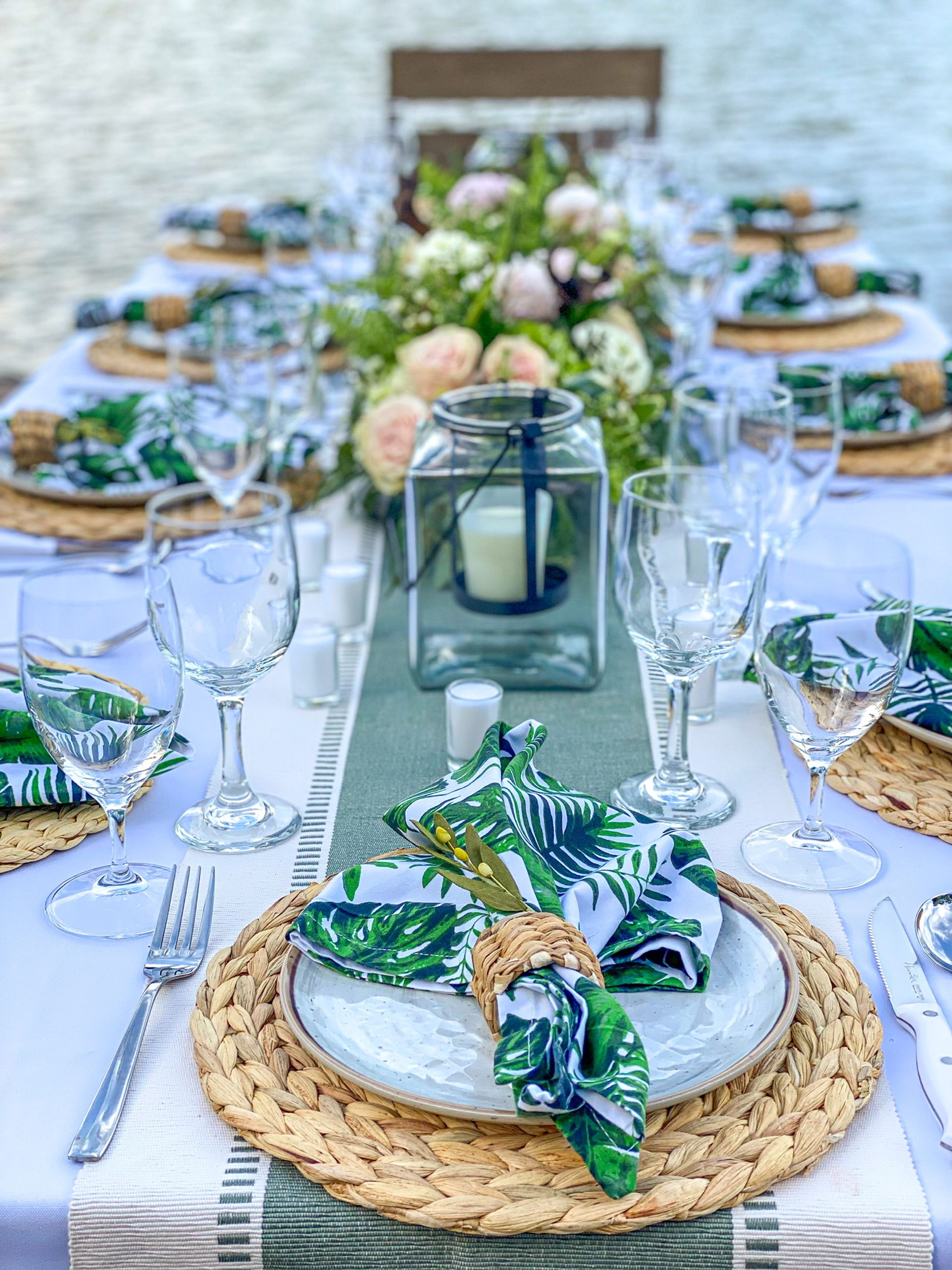Al fresco waterfront tablescape with woven chargers, white plates, tropical leaf napkins in rattan rings, clear wine glasses, a glass lantern candle and a lush floral centerpiece.