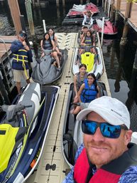 Group of people in life jackets on jet skis lined up along a floating dock at a marina, man in a white cap and mirrored sunglasses taking a selfie in the foreground — sunny waterfront adventure vibe