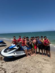 Friends in life jackets posing on a sunny sandy beach beside a white-and-blue jet ski with calm blue ocean and clear sky