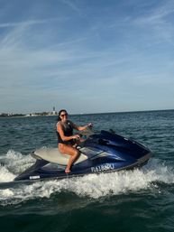 Smiling rider in a life vest on a blue jet ski cruising along the Florida coast with a distant lighthouse and clear blue sky.