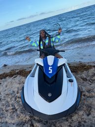Person with long braids wearing a neon striped swim top posing on a blue-and-white jet ski numbered 5 at a sandy beach with ocean waves in the background.