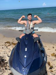 Smiling man in a life vest flexes his arms while sitting on a blue jet ski on a sunny sandy beach with gentle turquoise ocean waves.