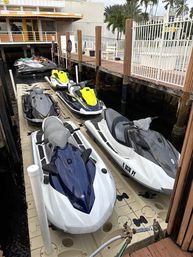 Row of colorful jet skis (personal watercraft) lined up on a floating dock beside a wooden pier and white fence at a waterfront marina with palm trees and a yellow‑awned building.