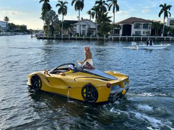 Yellow amphibious sports car cruising a palm-lined waterfront canal with a driver and a standing passenger waving a small American flag, luxury homes and a small boat in the sunlit background.