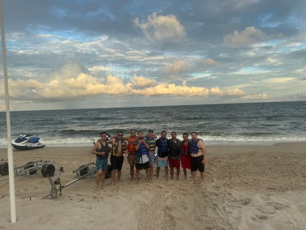 Group of people in life jackets posing on a sandy beach by the ocean at golden hour, dramatic cloud-filled sky overhead, with a parked jet ski and trailers on the shore.