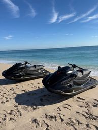 Two sleek black jet skis parked on a sunlit sandy beach next to calm turquoise ocean under a clear blue sky with wispy clouds