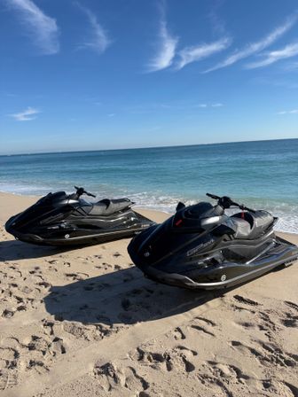 Two sleek black jet skis parked on a sunlit sandy beach next to calm turquoise ocean under a clear blue sky with wispy clouds