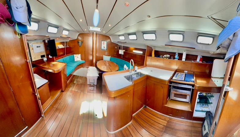 Bright cruising sailboat interior with warm wood paneling and teak floors, compact galley with double sink and stove, cozy turquoise U-shaped settee around a foldaway dinette, porthole windows and built-in storage.