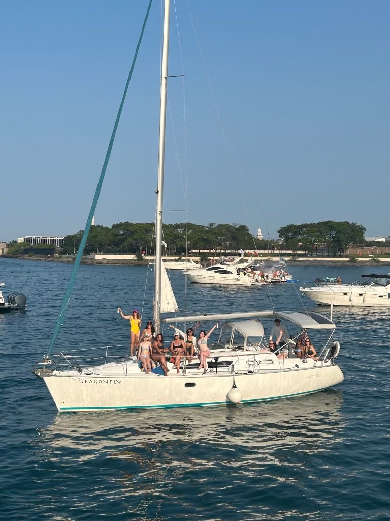 Sailboat party with a group of people in swimwear waving and lounging under a clear blue sky in a busy coastal harbor, surrounded by other yachts and a tree-lined shoreline.
