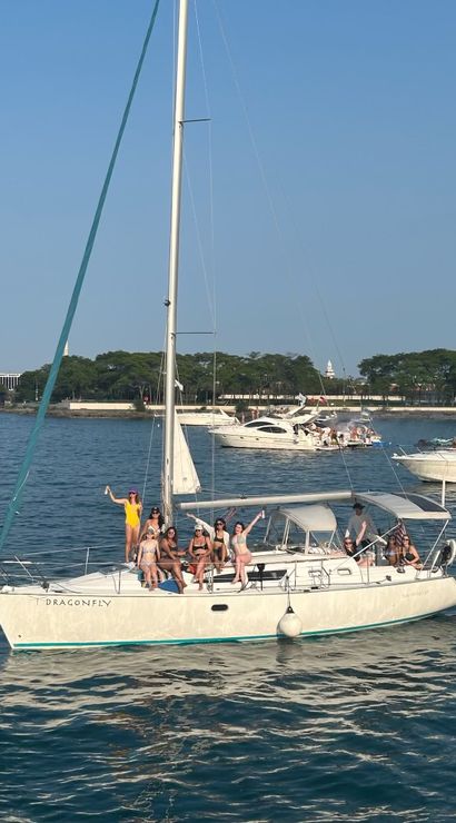 Sailboat party with a group of people in swimwear waving and lounging under a clear blue sky in a busy coastal harbor, surrounded by other yachts and a tree-lined shoreline.