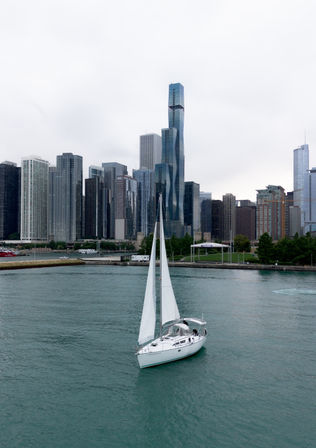 White sailboat gliding on turquoise lakefront waters with the Chicago skyline of modern skyscrapers under an overcast sky