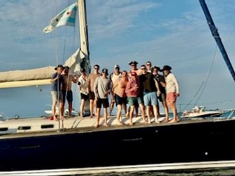 Group of friends posing on a sailboat deck, enjoying a sunny day on coastal waters with mast and flag against a blue sky