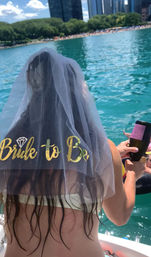 Woman wearing a gold-lettered “Bride to Be” veil on a boat, holding a bottle above turquoise water with a tree-lined beach and city skyline in the background — summer bachelorette on an urban waterfront.