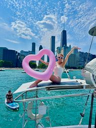 Person in a white swimsuit on a sailboat holding a pink heart-shaped float and wearing heart sunglasses, with turquoise Lake Michigan, a paddleboarder nearby and the Chicago skyline under a bright summer sky.