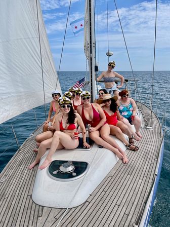 Cheerful group of women in swimsuits and captain hats lounging on the bow of a sailboat with sails up, flags flying and blue sea on the horizon.