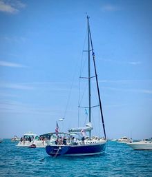 Blue-hulled sailboat with tall mast anchored among several yachts in clear turquoise coastal waters, groups of people on deck enjoying a sunny summer boating day